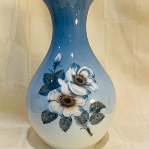 Peut inclure: Un vase en céramique bleu et blanc avec un motif floral. Le vase présente un dégradé de bleu à blanc et des fleurs blanches avec des centres bruns et des feuilles vertes.