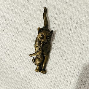 May include: A bronze cat figurine depicting a mother cat carrying a kitten. The cat has detailed features, including visible eyes and fur texture. The kitten hangs from the mother cat's mouth. The figurine is set against a textured, off-white background.