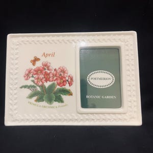 May include: A rectangular white ceramic picture frame featuring a raised leaf border. The left side displays a botanical illustration of pink Primula Obconica flowers, a butterfly, and the text April. The right side has a green insert with Portmeirion Botanic Garden text.