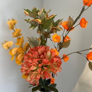 May include: A white ceramic jug displays a colourful floral arrangement. The arrangement includes a large, multi-layered orange and green flower, with yellow and orange accents. Green and brown leaves complete the natural look.