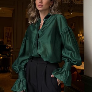 May include: A woman's fashion ensemble featuring a dark green silk blouse with long, ruffled sleeves and black high-waisted trousers. A small, dark gray handbag is held in the left hand. The setting appears to be indoors, possibly a restaurant or cafe.