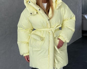 May include: A pale yellow puffer jacket with a large collar, button closures, and a matching belt. The jacket has large pockets and a quilted design. The model is wearing a white shirt underneath.