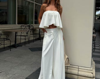 May include: A white two-piece outfit consisting of a strapless top and wide-leg pants. The top has a ruffled design, and the pants are high-waisted. A beige handbag is carried.
