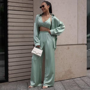 May include: A mint green satin three-piece outfit, including a crop top, wide-leg pants, and a button-down shirt. The outfit is paired with a white handbag and sandals. The model is wearing sunglasses.
