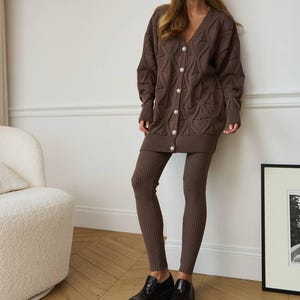 Op de afbeelding: Een bruine gebreide set bestaande uit een cardigan met lange mouwen en parelmoer knopen en bijpassende legging. De cardigan heeft een V-hals en een textuurpatroon. De outfit wordt gecombineerd met zwarte lakleren schoenen. Geschikt voor casual wear.