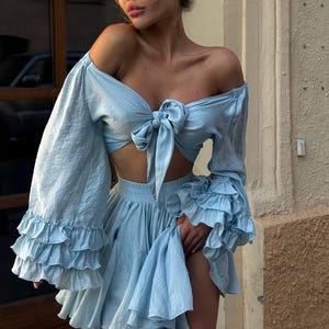 Könnte beinhalten: Hellblaues zweiteiliges Outfit mit einem bauchfreien Oberteil mit langen, gerüschten Ärmeln und einer Schleife an der Brust. Der passende Rock hat einen gerüschten Saum. Das Outfit ist aus einem weichen, fließenden Stoff.