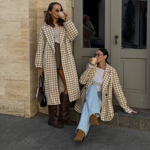 May include: Two people wearing houndstooth patterned coats and drinking from paper cups. One person is standing, wearing brown boots, a skirt, and carrying a black purse. The other person is sitting, wearing blue jeans and brown boots.
