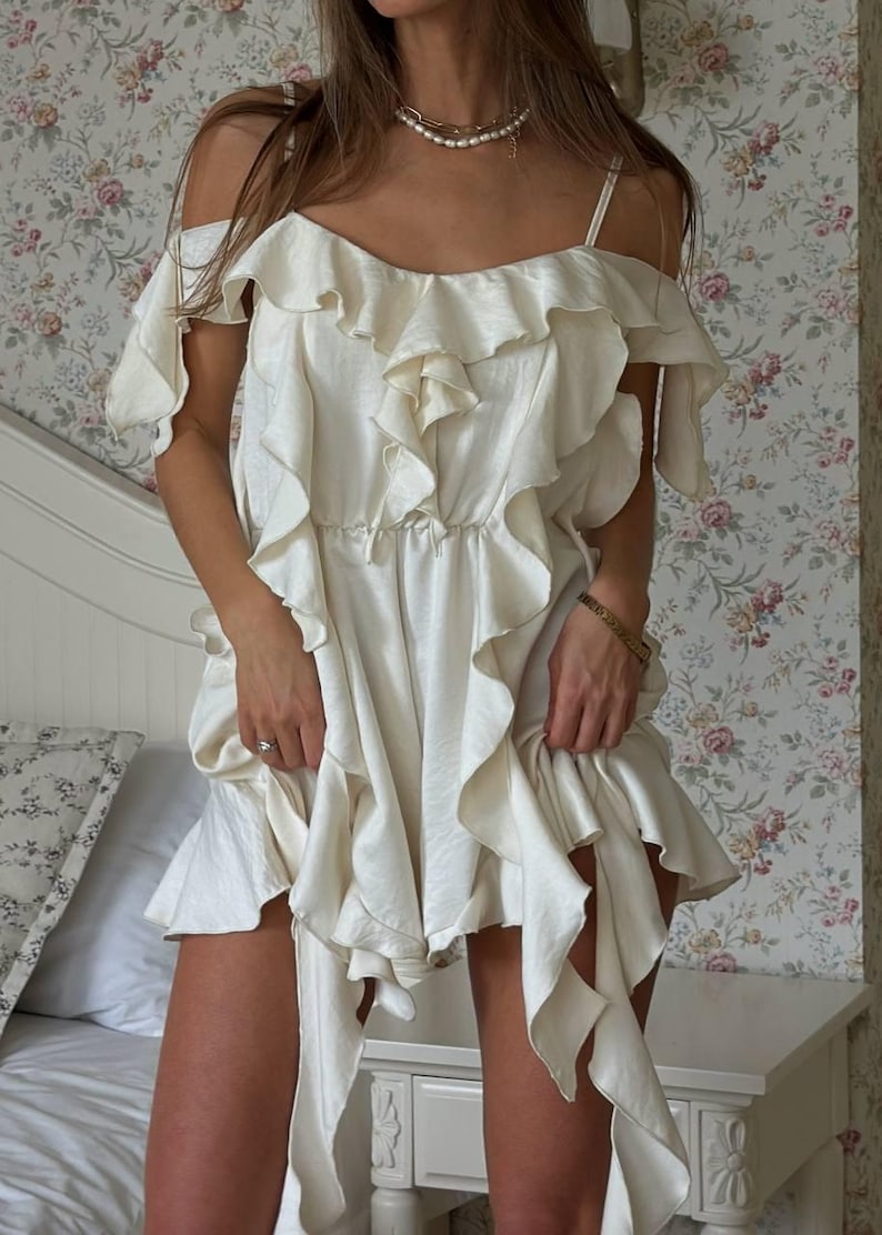 May include: Cream-coloured, ruffled dress with spaghetti straps and off-the-shoulder sleeves. The dress has multiple layers of ruffles and a cinched waist. The model is wearing a gold bracelet and a pearl necklace. The background is a floral wallpaper.