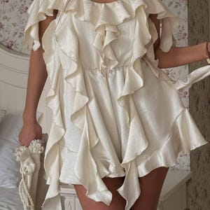 May include: Cream-coloured, ruffled romper with spaghetti straps and off-the-shoulder sleeves. The romper has multiple layers of cascading ruffles and a cinched waist. A matching cream bag with a decorative handle is held in the hand.
