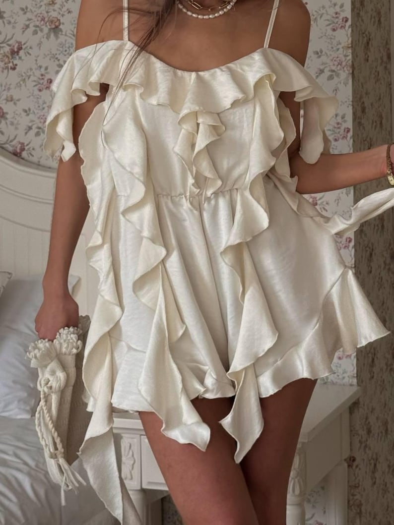 May include: Cream-coloured, ruffled romper with spaghetti straps and off-the-shoulder sleeves. The romper has multiple layers of cascading ruffles and a cinched waist. A matching cream bag with a decorative handle is held in the hand.