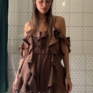 May include: A brown, ruffled mini dress with spaghetti straps and off-the-shoulder ruffles. The dress has a flowing, layered design and is made of a silky material. The model is wearing a gold bracelet and necklace.