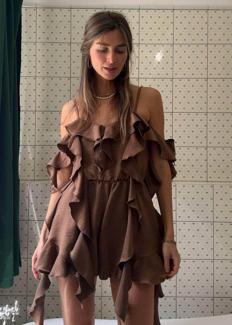 May include: A brown, ruffled mini dress with spaghetti straps and off-the-shoulder ruffles. The dress has a flowing, layered design and is made of a silky material. The model is wearing a gold bracelet and necklace.