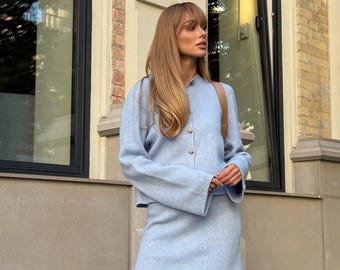 May include: A light blue knit two-piece set consisting of a long-sleeved button-down cardigan with a round neckline and a matching mini skirt. The cardigan features visible buttons down the front and ribbed cuffs, while the skirt has a simple hem.