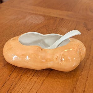 May include: A ceramic salt cellar with a light orange exterior and a white interior. A small white spoon rests inside the cellar. The salt cellar is oval-shaped and sits on a wooden surface.