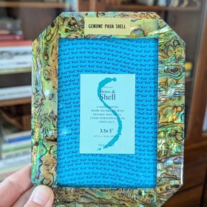 May include: A handcrafted photo frame with a genuine paua shell border. The frame has a blue interior and a label that reads "Natural Brass & Shell". The frame measures 3.5 x 5 inches.