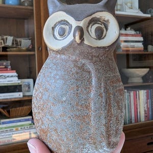 May include: A ceramic owl figurine with a brown and gray speckled glaze. The owl has large, round eyes with black pupils and white irises.