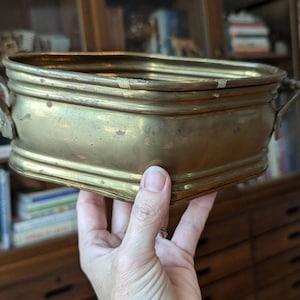 May include: A brass planter with two handles. The planter is oval shaped and has a worn, aged look.