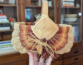May include: Handwoven basket with a natural, beige base and a decorative front panel in shades of orange and burgundy. The basket features a wooden handle and a geometric design on the front. A small tassel and wooden heart add detail.