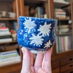 May include: A handmade ceramic cup with a blue glaze and white starburst designs. The cup has a rounded shape and a textured surface, showcasing the artistry of the pottery.