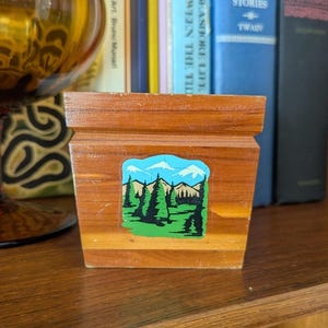 May include: A small, wooden box with a landscape sticker featuring green trees, blue mountains, and a blue sky. The box is made of light brown wood and sits on a wooden shelf. A vintage glass and books are in the background.
