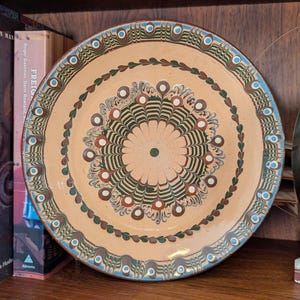 May include: A large, round, decorative ceramic plate with a hand-painted design. The plate features a beige background with concentric rings of detailed patterns in green, red, and blue. The plate is approximately 12 inches in diameter.