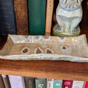 May include: A rectangular ceramic tray with a light brown and white speckled glaze. The tray has a series of brown, heart-shaped designs along the top edge. The tray is displayed on a wooden shelf, with books and decorative items in the background.