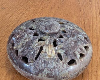 May include: A carved stone incense burner with a lid. The burner is oval-shaped and features intricate floral and leaf designs with cut-out patterns. The stone has a mottled appearance with shades of purple, gray, and tan.