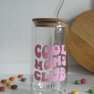 May include: A clear glass tumbler with a bamboo lid and a glass straw. The tumbler features the text "COOL MOMS CLUB" in pink lettering. Colorful candies are scattered around the tumbler.