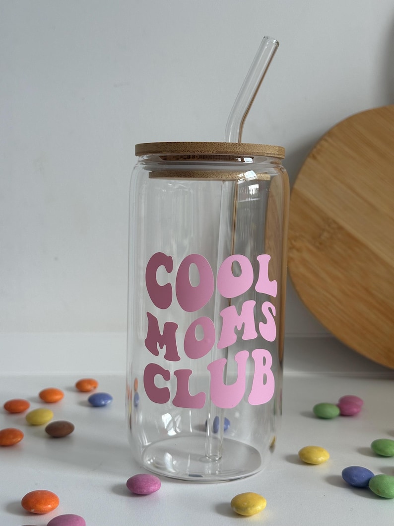 May include: A clear glass tumbler with a bamboo lid and a glass straw. The tumbler features the text "COOL MOMS CLUB" in pink lettering. Colorful candies are scattered around the tumbler.