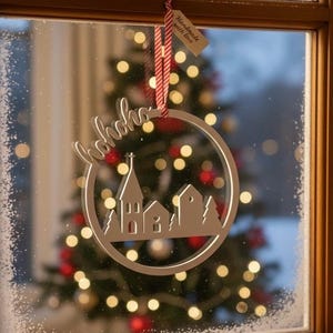 Könnte beinhalten: Silberner Weihnachtsschmuck mit einem kreisförmigen Rahmen und einem ausgeschnittenen Design einer Dorflandschaft, einschließlich Häusern und Bäumen. Das Wort "hohoho" ist unten ausgeschnitten. Ein Anhänger mit der Aufschrift "Handmade with love" hängt oben.