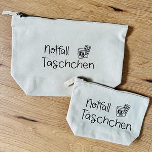 May include: Two off-white canvas zipper pouches. The larger pouch has a black zipper and the smaller pouch has a white zipper. Both pouches have the words "Notfall T&auml;schchen" and a medical symbol printed in black.