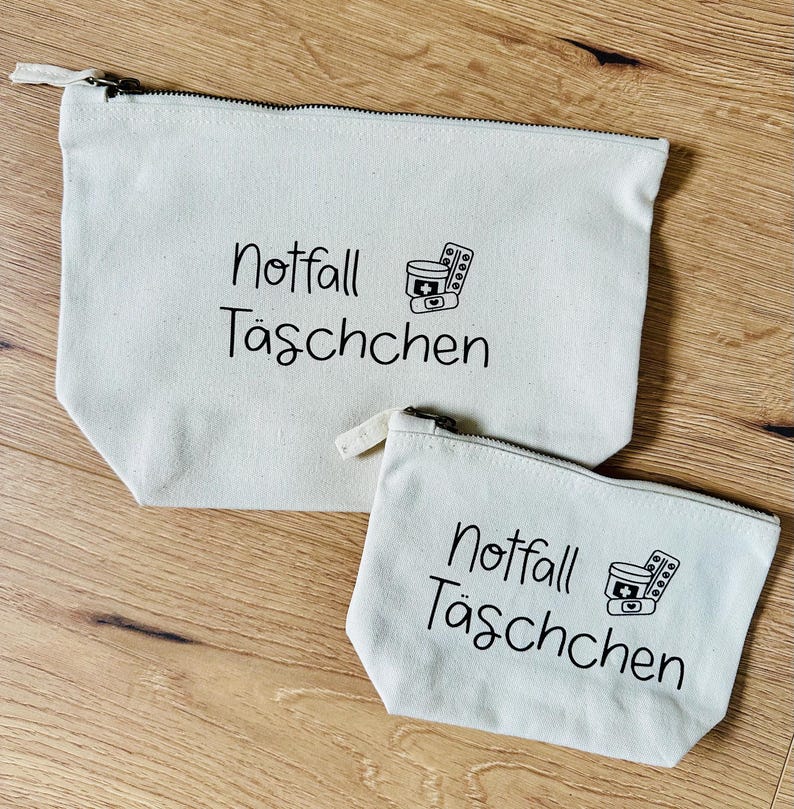 May include: Two off-white canvas zipper pouches. The larger pouch has a black zipper and the smaller pouch has a white zipper. Both pouches have the words "Notfall T&auml;schchen" and a medical symbol printed in black.