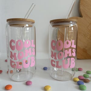 May include: Two clear glass tumblers with bamboo lids and glass straws. Each tumbler has the words "COOL MOMS CLUB" in pink lettering. Colorful candies are scattered around the tumblers.