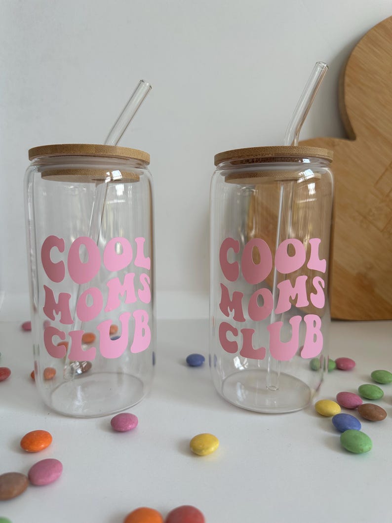 May include: Two clear glass tumblers with bamboo lids and glass straws. Each tumbler has the words "COOL MOMS CLUB" in pink lettering. Colorful candies are scattered around the tumblers.