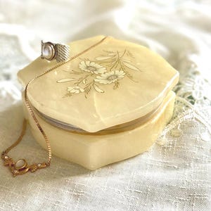 May include: A pale yellow, hexagonal stone trinket box with a smooth, polished finish and a delicate floral engraving on the lid. A silver-toned ring with a central stone and a gold-toned chain necklace with a small pendant rest on the box.