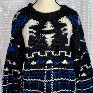 May include: A black, white, and blue knit jumper featuring a prominent graphic of two white polar bears facing each other near the neckline, with a geometric pattern of horizontal stripes and a plaid-like grid design across the lower body.