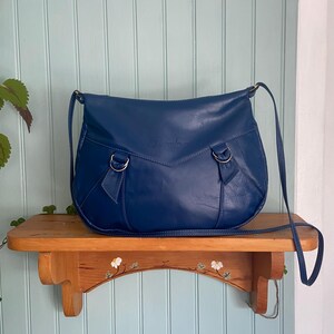 Vintage navy blue leather crossbody bag by Isadora on wooden shelf, crescent shape, thin strap, D-ring details, white green background