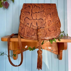 May include: A brown and tan snakeskin patterned leather shoulder bag with a fold-over flap closure. The bag features a central braided leather cord with a knotted detail and a long, dangling leather tassel. It includes a thin leather shoulder strap.
