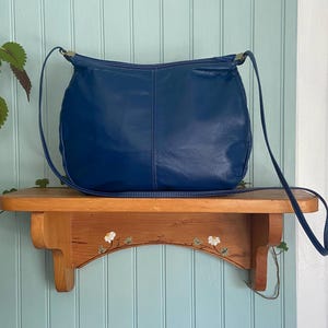 May include: A deep blue leather shoulder bag with a long strap. The bag is sitting on a wooden shelf with a floral design. The background is a light blue wall.