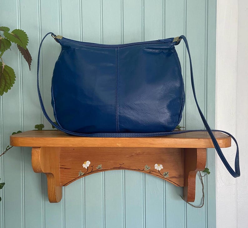 May include: A deep blue leather shoulder bag with a long strap. The bag is sitting on a wooden shelf with a floral design. The background is a light blue wall.