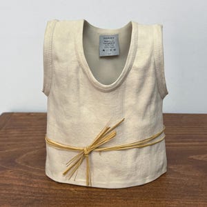 May include: A cream-coloured ceramic sculpture shaped like a sleeveless tank top with a ribbed texture. A label inside the neckline reads HARVEY, Made in CA, 100% Clay, 100% Argile. A thin, light brown raffia string is tied around the waist area.