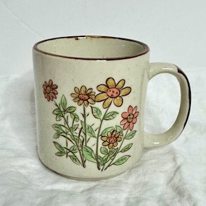 May include: Cream ceramic mug featuring a floral pattern of yellow and pink flowers with green leaves. The rim and handle are brown. A delightful mug for tea or coffee, perfect for the kitchen.