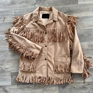 May include: A tan jacket featuring long fringe detailing across the chest, sleeves, and hem. The jacket includes a collared neckline, a button-front closure, and two front pockets with fringe trim. A black label is visible inside the collar.