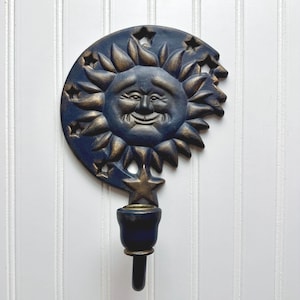 May include: A decorative wall sconce featuring a smiling sun face with a crescent moon and star accents. The sun and moon are dark blue with gold highlights. The sconce has a candle holder at the bottom.
