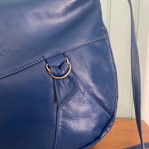 May include: A deep blue leather shoulder bag with a long strap. The bag features white stitching and a gold-colored metal ring. The word "Lora" is embossed on the front of the bag.