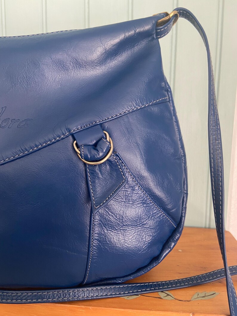 May include: A deep blue leather shoulder bag with a long strap. The bag features white stitching and a gold-colored metal ring. The word "Lora" is embossed on the front of the bag.