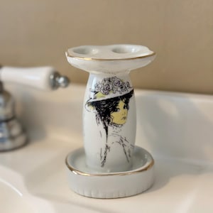 May include: White ceramic toothbrush holder with a gold rim and a decorative illustration of a woman in a hat. The holder has two toothbrush slots and a small base. The woman's portrait is in black, yellow, and white.