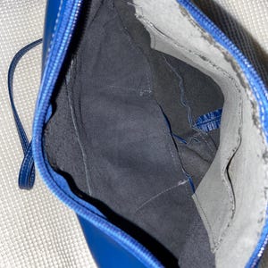 May include: Open view of a blue leather handbag with a zipper closure and a thin strap. The interior is lined with dark gray fabric and features pockets. The bag's stitching is white, and the overall design is sleek and modern.