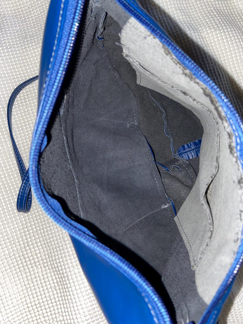 May include: Open view of a blue leather handbag with a zipper closure and a thin strap. The interior is lined with dark gray fabric and features pockets. The bag's stitching is white, and the overall design is sleek and modern.