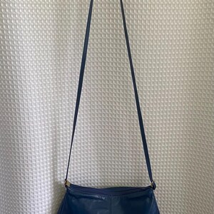 May include: A navy blue leather shoulder bag with a long strap. The bag has a rounded shape and a gold-colored clasp. The bag is hanging against a white textured background.